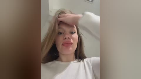 Video of a young Caucasian woman with straight brown hair, wearing a white top, making a silly face with her hand on her head, blurred background.