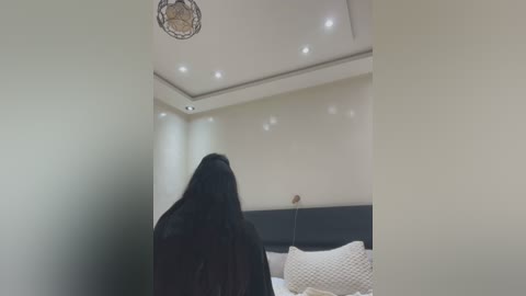 Video of a dark-haired woman with a long, straight mane, standing with her back to the camera in a minimalist bedroom. The room features a beige wall, recessed lighting, a dark headboard, and a textured, cream-colored bedspread.