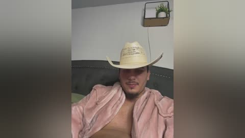 Media: Video of a fit, light-skinned man with medium-length dark hair, wearing a cowboy hat and pink robe, sitting on a grey bed with a green pillow. The background features a minimalist wall and a small plant in a wooden shelf.