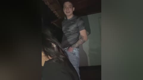 Media: Video of a muscular, tattooed man with short hair in a black striped shirt and jeans, smiling in a dimly lit room with dark, blurry figures.