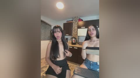 Video of two young women with long hair, one topless, in a modern kitchen; one with a computer on her lap.