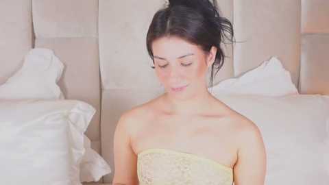 Video of a slender, fair-skinned woman with dark hair in a messy bun, wearing a strapless yellow lace dress, sitting on a white bed in a softly lit, minimalist bedroom.