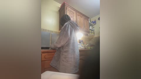 Media: A video of a person with shoulder-length dark hair, wearing a gray hoodie, standing in a dimly lit kitchen with wooden cabinets, a sink, and a tiled backsplash.