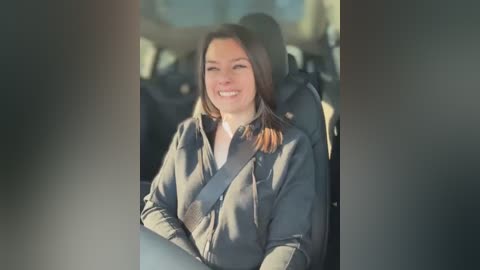 Video of a smiling woman with shoulder-length brown hair, wearing a gray jacket, seated in a car with a sunlit window background.