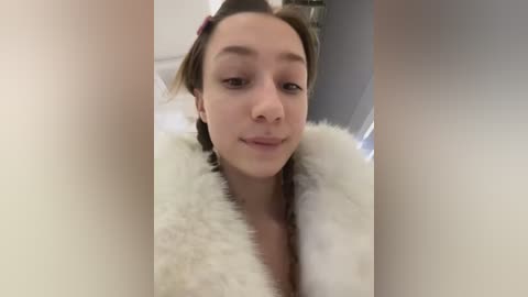 A video of a young woman with light skin and dark hair, wearing a white fur coat, taken indoors with a blurred background.