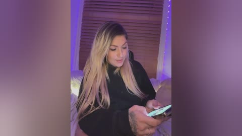 Media: Video of a young, light-skinned woman with long, straight blonde hair, wearing a black sweater, sitting on a bed with a fur blanket, holding a smartphone.