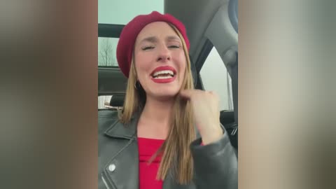 Media: Video of a smiling woman with long blonde hair and fair skin, wearing a red beret, grey leather jacket, and red top, seated in a car, with blurry out-of-focus people in the foreground.