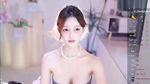 Video of a young East Asian woman with fair skin, brown hair, wearing a strapless dress and pearl necklace, sitting in a modern room with a blurred background.