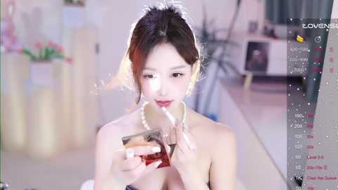 Video of an Asian woman with fair skin and dark hair, wearing a pearl necklace, drinking from a glass of red liquid, set in a softly lit, minimalist room.