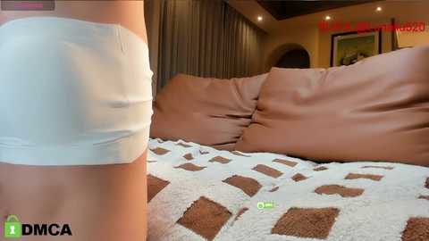 Media: Video of a woman in a white mini skirt lying on a bed with brown and beige patterned blanket, surrounded by beige pillows.