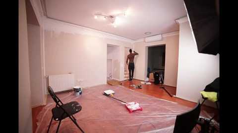 Media: Video of a partially painted room with a man painting the ceiling, white walls, a black folding chair, painting supplies scattered on the floor, and a black camera stand.