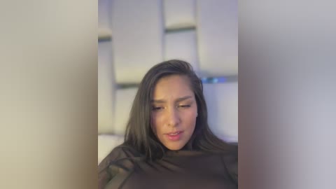 Media: Video of a Latina woman with medium skin tone, long dark hair, wearing a black top, looking down, in a modern, minimalist room with white tiles.