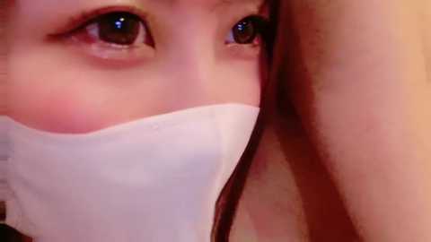 Video of a young Asian woman with light brown skin, large brown eyes, and a white surgical mask covering her nose and mouth. Her right arm is partially visible, and she is indoors with a warm, dimly lit background.