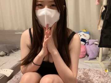 Media: Video of an East Asian woman with long brown hair, wearing a white face mask, black tank top, and black shorts, kneeling on a plush rug, clasping hands in prayer pose. Background includes a bed, stuffed animals, and a mirror.