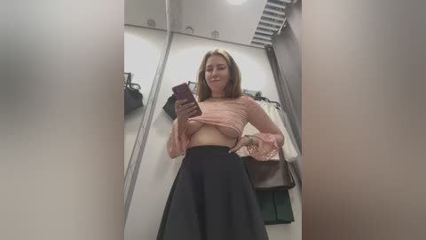 Video of a smiling woman with light skin and brown hair, wearing a pink crop top and black skirt, taking a selfie in a mirrored fitting room.