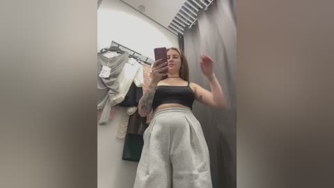 Video of a young, tattooed woman with light skin and brown hair, wearing a black crop top and high-waisted white pants, taking a mirror selfie in a clothing store.