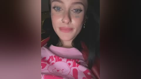 A video of a young woman with light skin, blue eyes, and dark hair, wearing a pink shirt with a bear design. She has a nose ring and a small blue tattoo on her cheek.