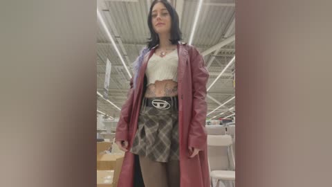 Video of a young woman with fair skin and dark hair, wearing a white lace crop top, a plaid skirt, and a long pink coat, standing in a modern warehouse with high ceilings and exposed pipes.