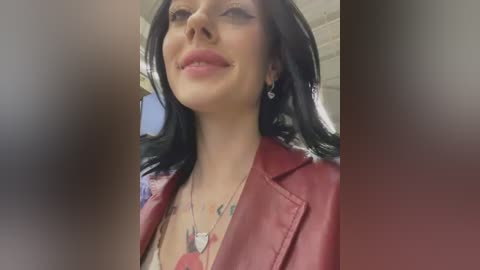 Video of a smiling woman with long black hair, wearing a red leather jacket, and a necklace with a heart pendant. She has a tattoo on her left chest and is indoors.