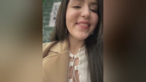 Media: Video of a young woman with long, straight brown hair, wearing a beige coat and white lace-up top, smiling slightly, with a blurred background of greenery.