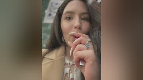 Media: Video of a young woman with long dark hair, light skin, and green eyes, biting her finger, wearing a beige jacket and a floral-patterned blouse, in a blurred, indoor setting.