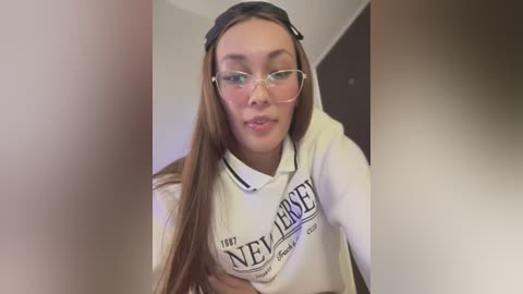 Video of a young woman with light skin, long straight brown hair, wearing glasses and a white sweatshirt with \"NEVER\" in bold letters. She's indoors with a blurred background.