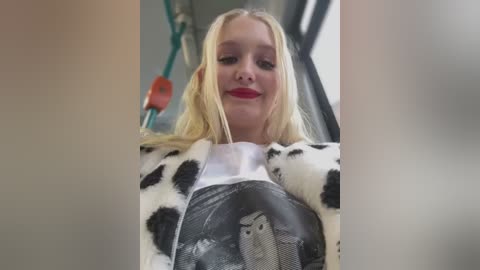 A video of a smiling young woman with straight blonde hair, wearing a white blouse with a black cow print, standing in a bus.