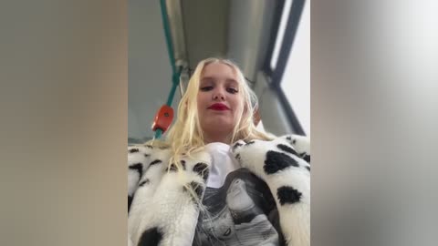 Video of a young blonde girl with fair skin and rosy cheeks, wearing a white shirt and black-and-white cow patterned jacket, sitting in a train with teal seats.