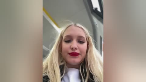 Video of a young blonde woman with long hair, red lipstick, wearing a black jacket, inside a bus. Background shows bus interior with yellow ceiling strip.