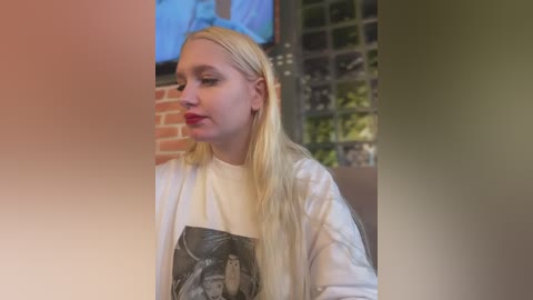 Video of a young blonde woman in a white shirt, standing indoors near a TV screen and a window with frosted glass.