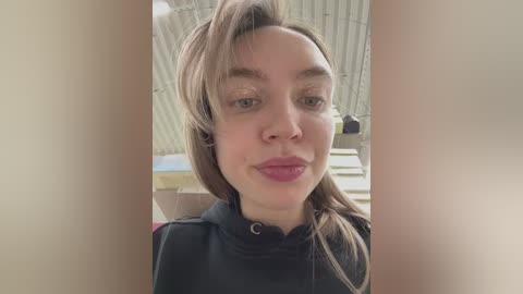 Media: Video of a Caucasian woman with fair skin, light brown hair, and subtle makeup, wearing a black top, taken indoors against a corrugated metal ceiling background.