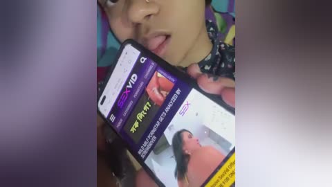 Media: Video of a young woman with a nose ring, wearing a colorful patterned headscarf, holding a smartphone displaying a video of a woman's face with a large mole on her cheek.