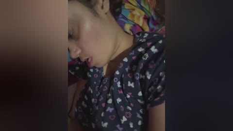 Media: Video of a young child with light brown skin, wearing a dark blue shirt with a pattern of hearts and butterflies, lying down with eyes closed, surrounded by a colorful, floral-patterned pillow.