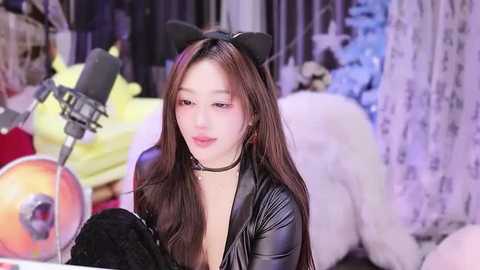 Video of an East Asian woman with long, straight brown hair, wearing a black choker, black cat ears, and a black leather jacket. She is singing into a microphone, surrounded by plush toys and a colorful background.