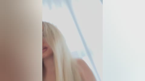 Media: Video of a blonde woman with long hair partially obscuring her face, standing in front of a tall, white vertical object, likely a window or pillar, in a softly lit room.