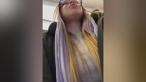 Video of a young woman with long blonde hair, wearing glasses, a gray shirt, and a black jacket, looking tired and leaning back in a car seat.