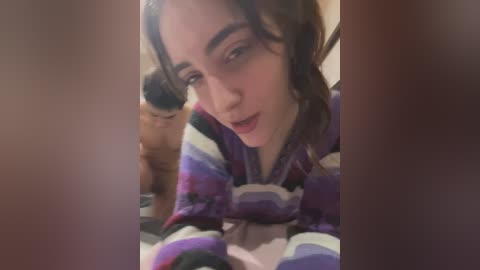 Media: A video of a young woman with wavy brown hair and a fair complexion, wearing a multicolored striped sweater, leaning forward with a playful expression. The background is blurry and dimly lit, making her the main focus.