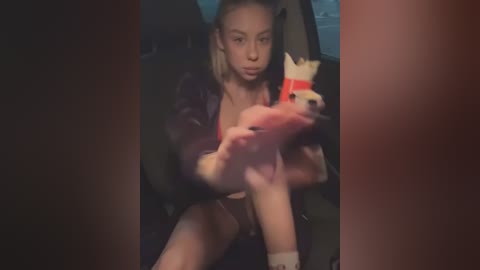 Video of a blonde woman with fair skin, wearing a black jacket, holding a plush toy with a red collar inside a dark car.