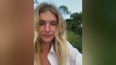 A video of a blonde woman with wavy hair and fair skin, wearing a white shirt, smiling. Background shows green foliage and a clear blue sky.