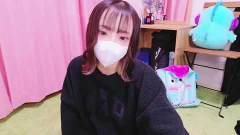 Media: Video of a young Asian woman with shoulder-length brown hair, wearing a black sweater, white face mask, and pinkish lipstick. Background features a pink curtain, stuffed unicorn toy, and green carpet.