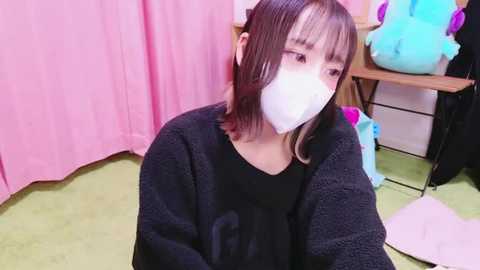 Media: Video of a young Asian woman with shoulder-length brown hair, wearing a black sweater and white mask, sitting on a green floor in a room with pink curtains, blue plush toys, and a wooden desk.