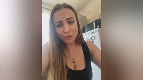 Media: A video of a woman with long brown hair, wearing a black tank top, standing in a kitchen with a microwave and refrigerator in the background. She has a neutral expression and is slightly out of focus.