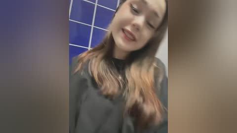 Video of a young woman with light skin and shoulder-length, wavy, auburn hair, smiling while wearing a black hair cape in a tiled bathroom with blue and white tiles.