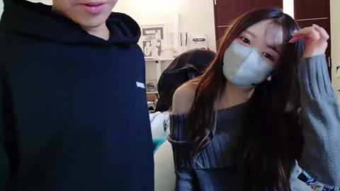 Media: Video of a young East Asian woman with long dark hair, wearing a gray off-shoulder top and a face mask, standing next to a man in a black hoodie in a dimly lit room with shelves in the background.