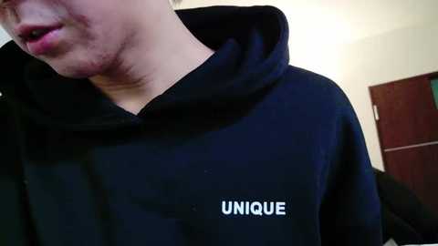 Media: Video of a person with light skin wearing a black hoodie with the word \"UNIQUE\" in white letters on the chest. The background shows a white wall and a wooden door. The image focuses on the person's head and upper torso.