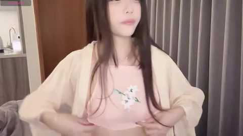 Media: Video of a young, pale-skinned woman with long black hair, wearing a light pink shirt and beige cardigan, holding her breasts. Background features a door and gray curtains.