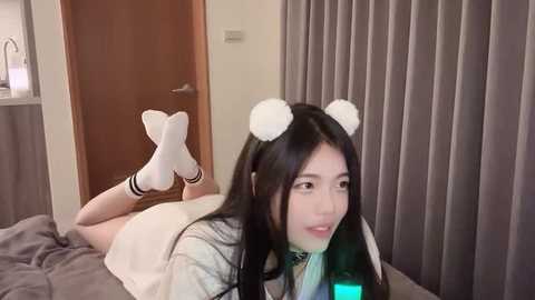 Media: Video of an Asian woman with long black hair, wearing white socks, a white skirt, and white earflap hat, lying on a bed with grey sheets and curtains, smiling.