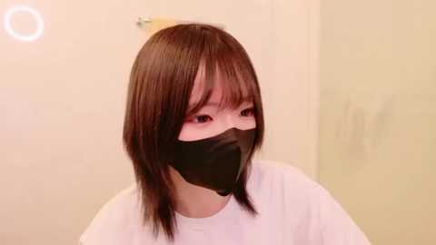 Media: Video of an East Asian woman with straight, shoulder-length brown hair and bangs, wearing a black face mask and a white top, against a plain beige wall.