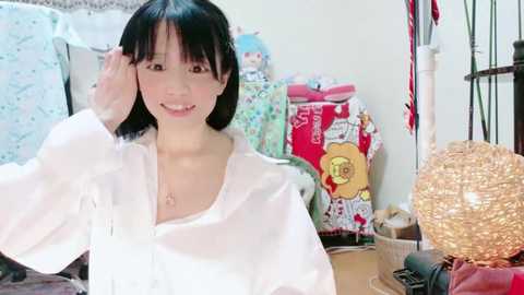 Video of a smiling Japanese woman with straight black hair, wearing a white shirt, in a cluttered room with colorful, patterned fabric and a spherical lamp.