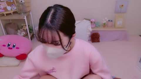 Video of a young woman with dark hair, wearing a pink sweatshirt and face mask, sitting in a pink room with Kirby plushies and a shelf with toys and decor.